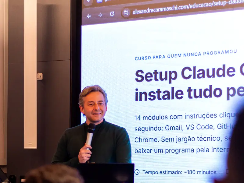 Alexandre Caramaschi, com um microfone, apresenta numa sala de treinamento a página do curso Setup Claude Code do portal; alunos aparecem em primeiro plano, fora de foco.